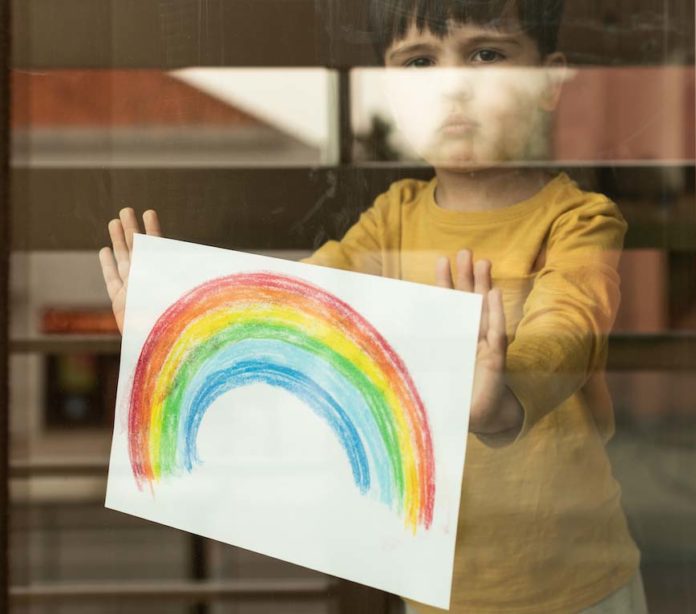 Little kid holding a drawing of a rainbow through the window