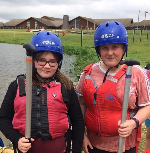 Two girls, wearing life jackets and blue helmets and carrying oars. 