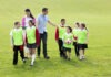 Learning outside the classroom in unprecedented times Group of school children wearing bibs and school uniform outside on the grass with their teacher. One of the boys is holding a soccor ball.