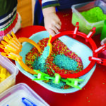 Toddlers playing with sensory bin with colourful rice on red table.