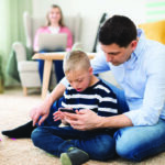 Father with happy down syndrome son indoors at home, using smartphone.